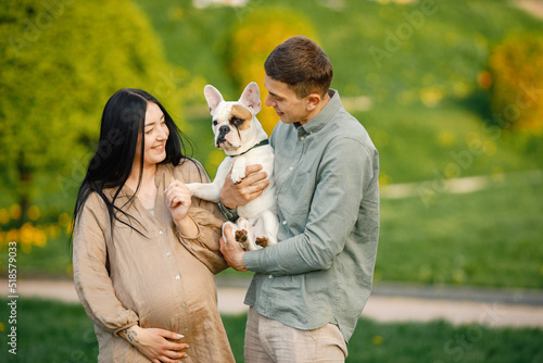 Pregnant woman and her husband standing in a park and holding little french bulldog on a hands © hetmanstock2