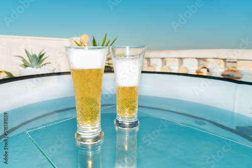 Glasses with cool beer with foam on a glass table above the swimming pool on the terrace of a tropical hotel on a hot summer day.cooling drink at pool party