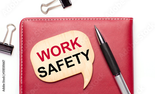 On a light background there are black paper clips, a pen, a burgundy notepad a wooden board with the text WORK SAFETY