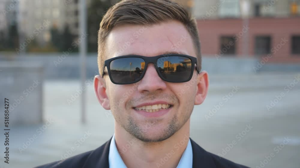Portrait of happy businessman in sunglasses with cityscape at background. Handsome business man looking at the camera and smiling. Face of cheerful guy. Blurred background. Slow motion Close up Portrait of happy businessman in sunglasses with cityscape at background. Handsome business man looking at the camera and smiling. Face of cheerful guy. Blurred background. Slow motion Close up
