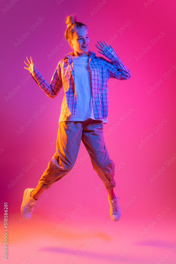 Jumping. Young beautiful girl, student in casual style clothes isolated on magenta color background in neon light. Concept of beauty, art, fashion, emotions