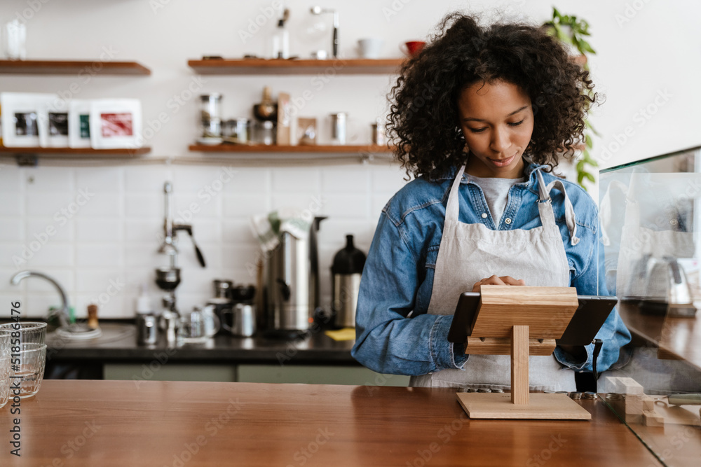 Black barista woman wearing apron working with tablet computer in cafe