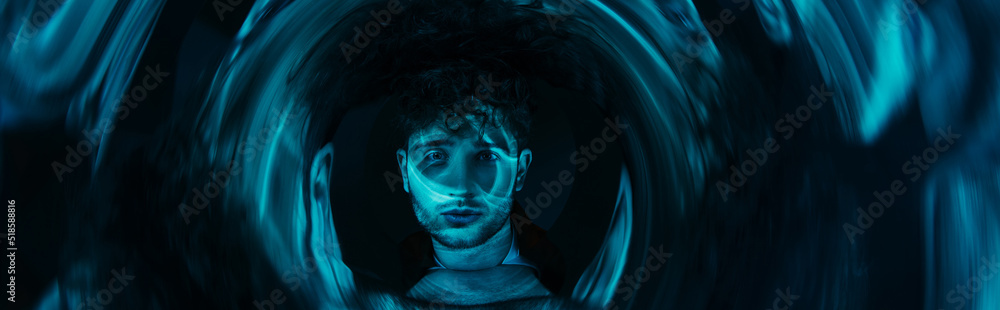 curly man looking at camera through futuristic neon blue circle on dark background, banner. curly man looking at camera through futuristic neon blue circle on dark background, banner.