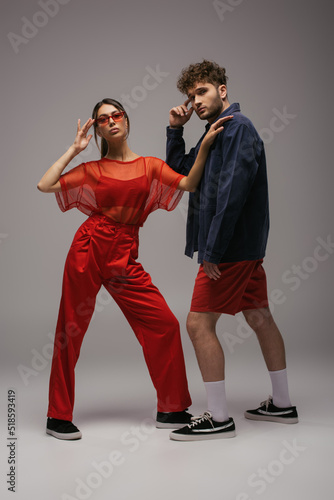 full length of stylish woman in red outfit and sunglasses posing near man on grey.