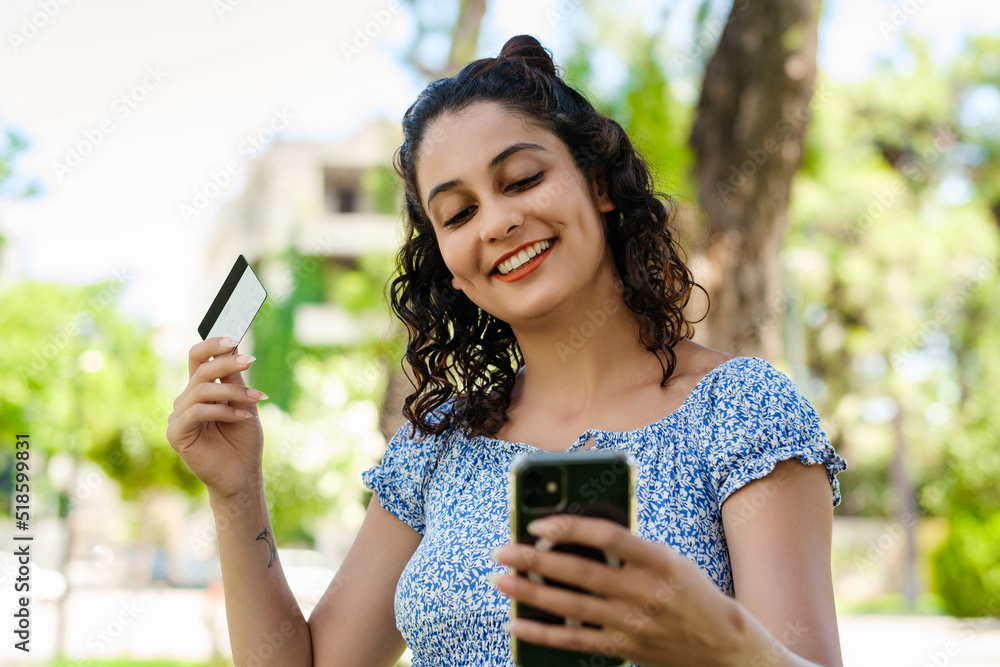 Cheerful brunette woman wearing summer dress on city park, outdoors holding mobile phone credit bank card do online shopping provide internet payment. Safe mobile banking.
