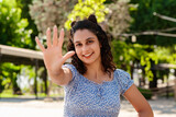 Young woman smiling confident wearing summer dress standing on city park, outdoors showing and pointing up with fingers number five while smiling confident and happy. Selective focus on her face.