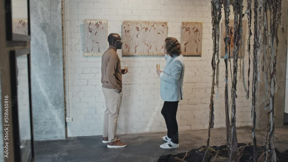 Long tracking right of young Caucasian man and his intelligent Black male friend holding champagne glasses, discussing series of paintings on exhibition opening at daytime Long tracking right of young Caucasian man and his intelligent Black male friend holding champagne glasses, discussing series of paintings on exhibition opening at daytime