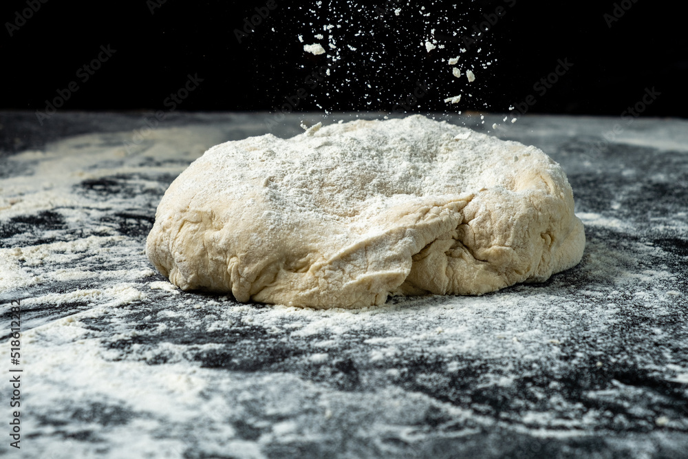 Flour is poured on fresh dough, dark background, lump of dough.