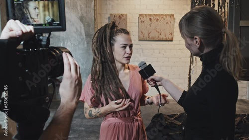 Medium of young Caucasian female artist with dreadlocks giving interview to journalist, unrecognizable cameraman filming in gallery on exhibition opening photo