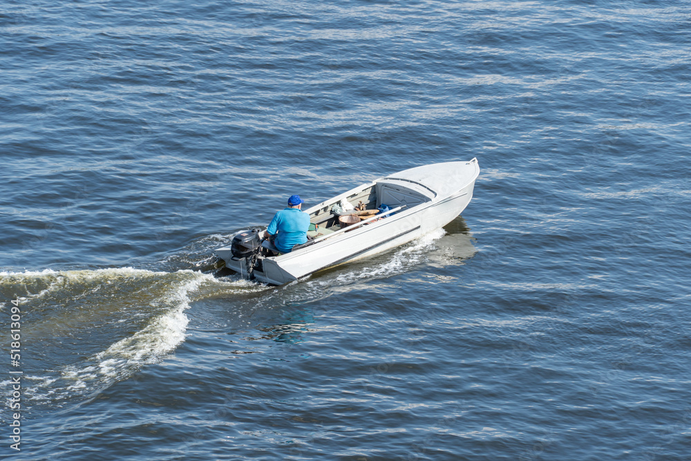 Fototapeta premium Adult man fast moving in white motorboat on the river. He drives the motor and looks for fish. Waves on the water and foam after speed. Water transport. Top view. Lifestyles.
