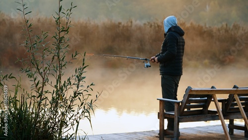 Fotografie, Obraz Hoodie male standing and fishing by fishing rod in the lake with grass and  plan