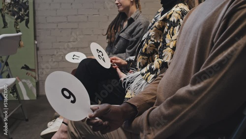 Midsection side-view of cropped buyers sitting on chairs indoors on auction, holding bid cards with numbers