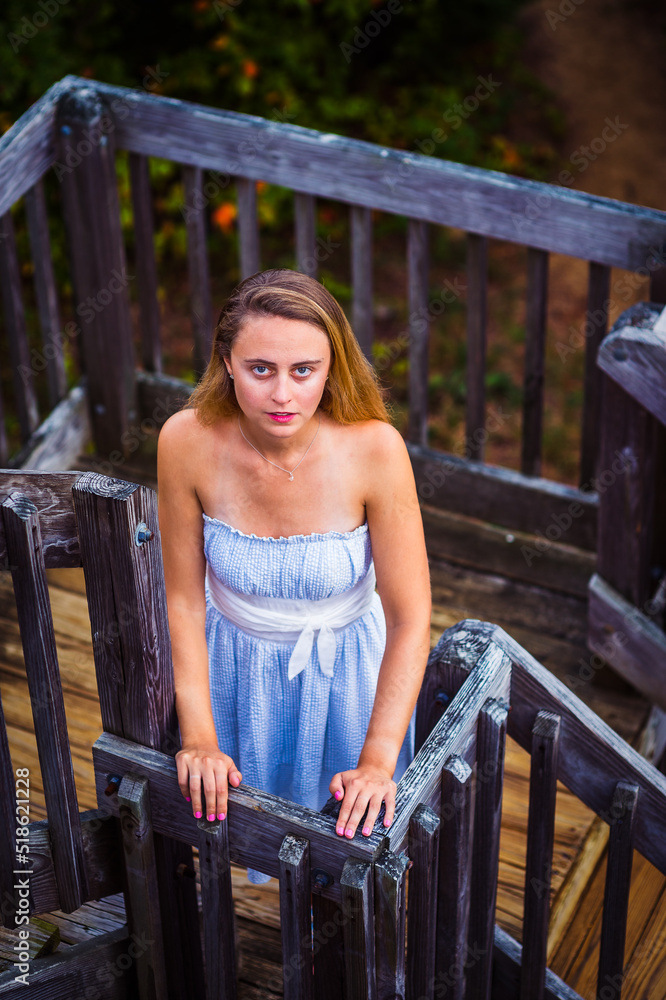 Looking Up. Young woman wearing white blue dress, white waistband, standing on wooden staircase, mysteriously looking up.