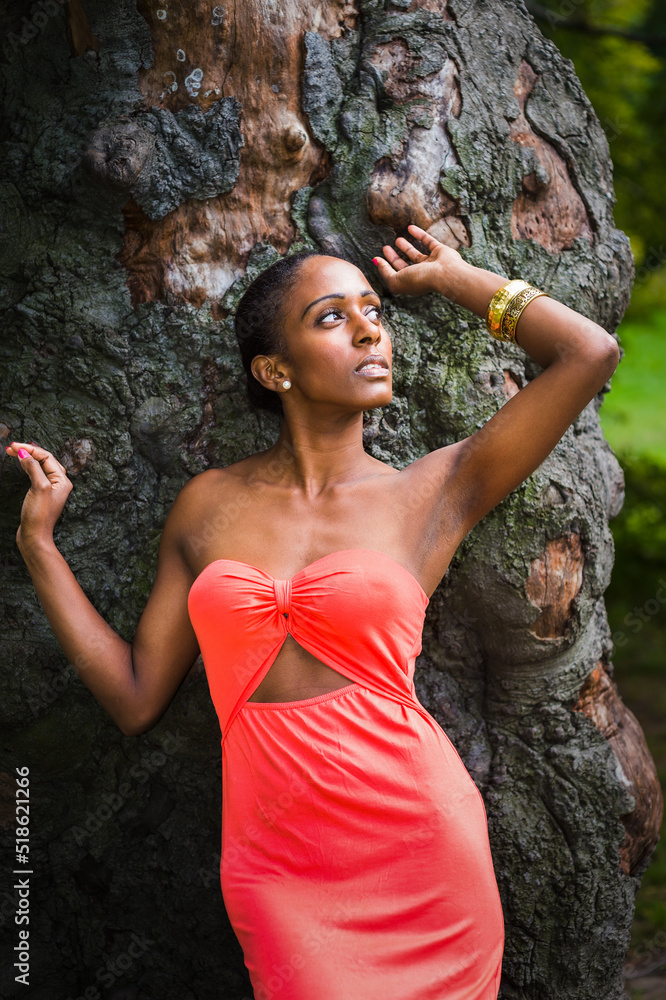 By the tree, Young woman wearing red two pieces dress, standing by old tree trunk, looking around, wondering.