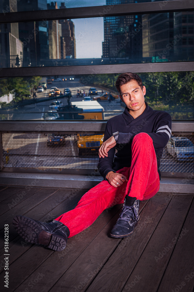 Young Man wearing black shawl collar sweater, red jeans, black leather shoes, sitting on floor by big glass wall in New York City, relaxing, thinking. Background is busy street scene.