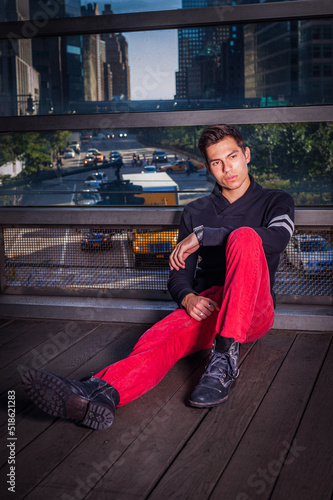Obraz na płótnie Young Man wearing black shawl collar sweater, red jeans, black leather shoes, sitting on floor by big glass wall in New York City, relaxing, thinking. Background is busy street scene. w salonie