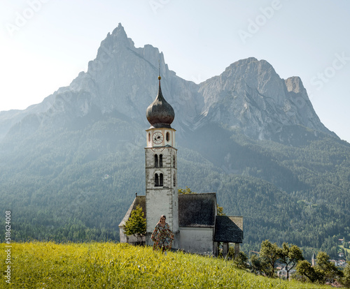 A woman traveler enjoys a visit to the church of San Valentino in the middle of a green valley surrounded by the mountains of the italian alps in dolomites