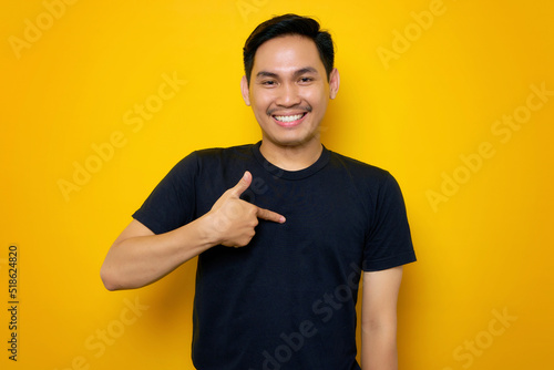 Cheerful young Asian man in casual t-shirt pointing at herself with proud isolated on yellow background. People lifestyle concept