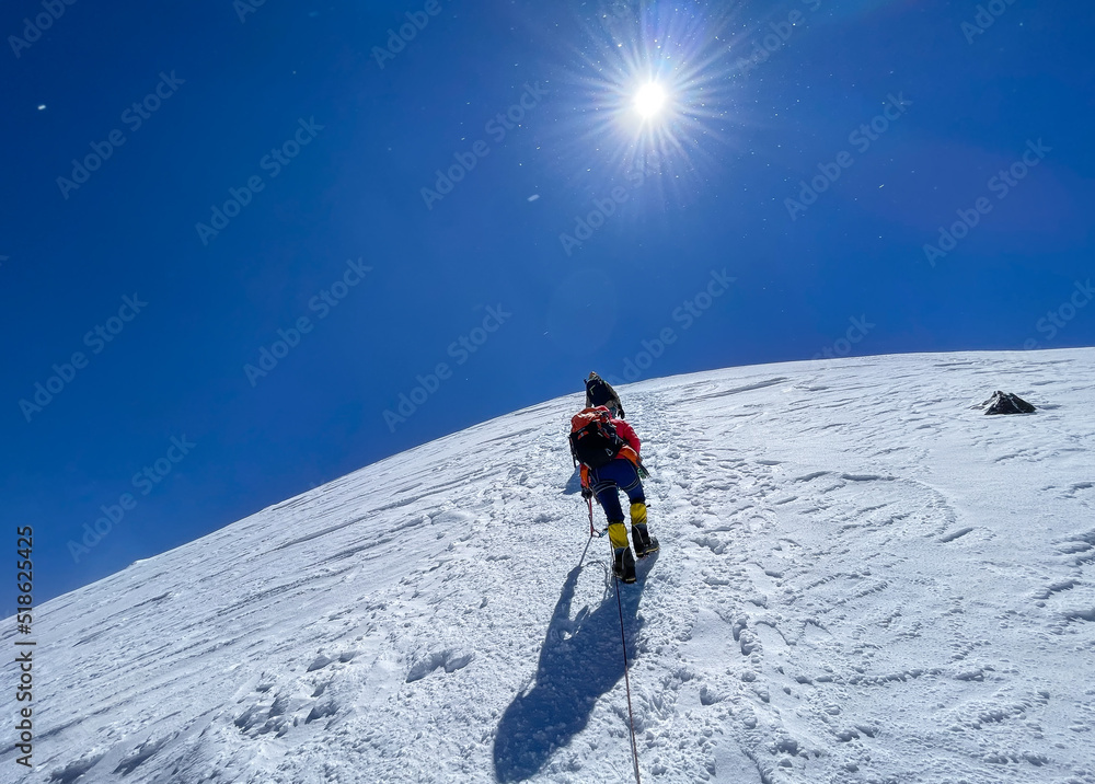 Last steps before Kazbek (Kazbegi) summit 5054m rope team dressed mountaineering clothes, boots with crampons ascending by snowy slope with blue sky background and bright sun. East Caucasus mountains.