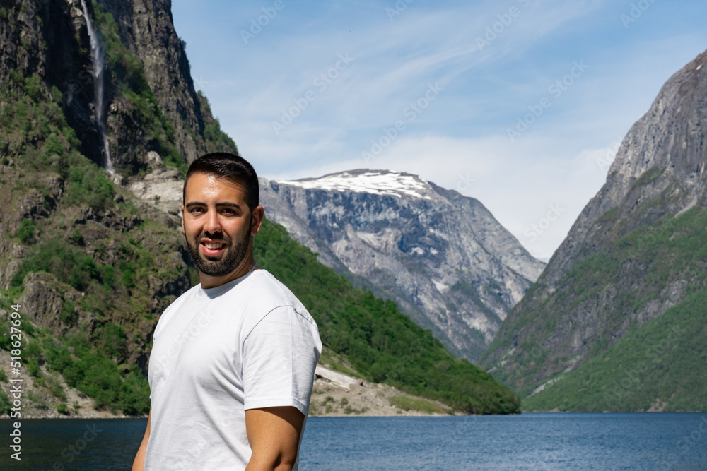 Young male tourist in the foreground smiling and behind him the high mountains and the fjord, in Gudvangen - Norway
