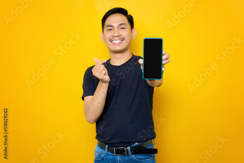 Cheerful young Asian man in casual t-shirt showing mobile phone with blank screen, showing korean heart gesture isolated on yellow background. People lifestyle concept