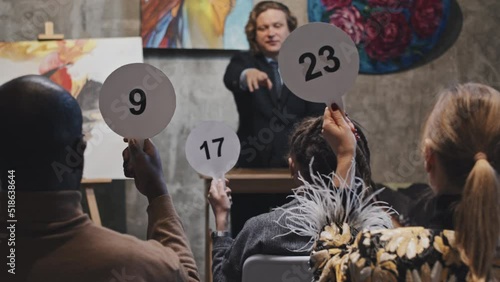 Medium long slow motion of young blond Caucasian man in formal suit leading auction and taking bids, rear of unrecognizable people raising up bidding cards