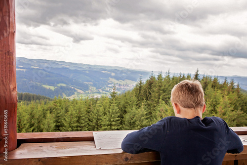 In the first plan young boy, child, on a watchtower. We see his back, he is reading, watching a map. In front of him is a mountain view, with pine forests and valleys. Rozhledna Milonova, czechia, EU photo