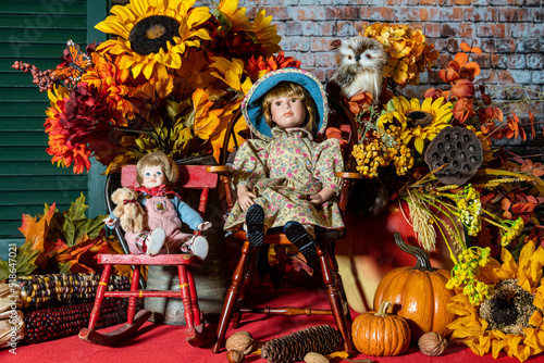 fall still life with children's dolls sitting on chairs surrounded by autumn foliage and pumpkins with owl and butterfly on red