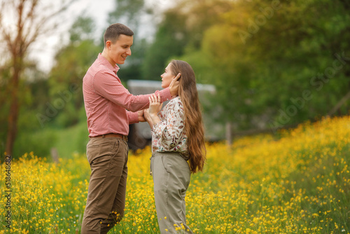 Young loving couple gently hugging, a lot of yellow flowers are around on the field. Enjoying time together. Man and woman are looking at each other.