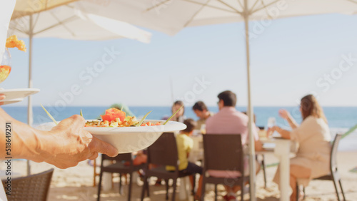 Professional waiter bringing different plates - paella, salad, prawns in tempura - to a family's table in a restaurant on the beach