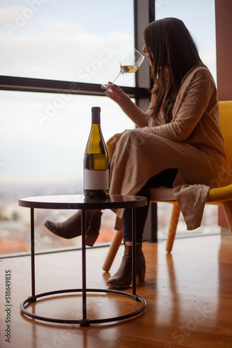woman sitting on the chair near low table with bottle of wine and drinking white wine and looking in window