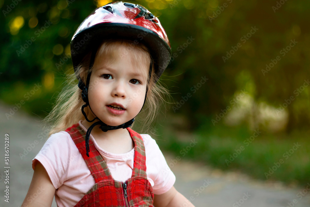 Close-up of face of cute little girl in red bicycle helmet looking at camera riding in the park in summer Close-up of face of cute little girl in red bicycle helmet looking at camera riding in the park in summer