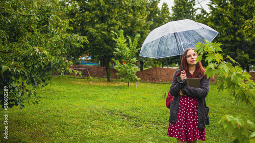 Young beautiful woman with transparent umbrella hugging book, standing in city park in rain. Dreamy female relaxing in nature with book, sheltering with umbrella from rain on walk in cloudy weather.