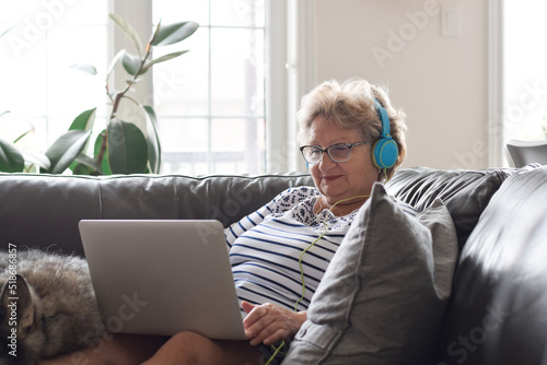 Fotografiet Senior woman in headphones sitting on couch at home using laptop computer