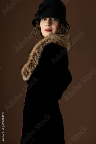 A 1920s woman wearing a black velvet coat with a fur collar and standing against a plain studio backdrop