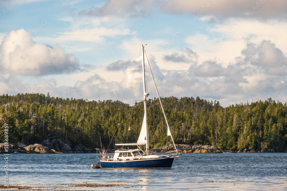 A sail boat cruising the Inland Passage at anchor in a sheltered bay on B.C.'s Central Coast, in Heiltsuk Territory.  Shot from the beach at mid day with white puffy clouds.