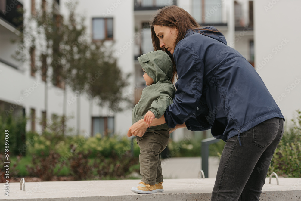A mother is checking the clothes of her pretty daughter in a raincoat on a cloudy day. A mom is teaching her female toddler how to do steps in the street at noon.
