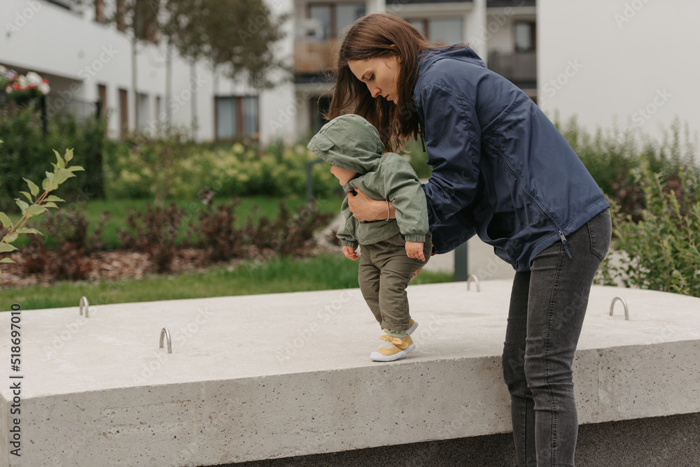 A happy mother is holding her young daughter in a raincoat on a cloudy day. A mom is teaching her female toddler how to do steps in the street at noon.