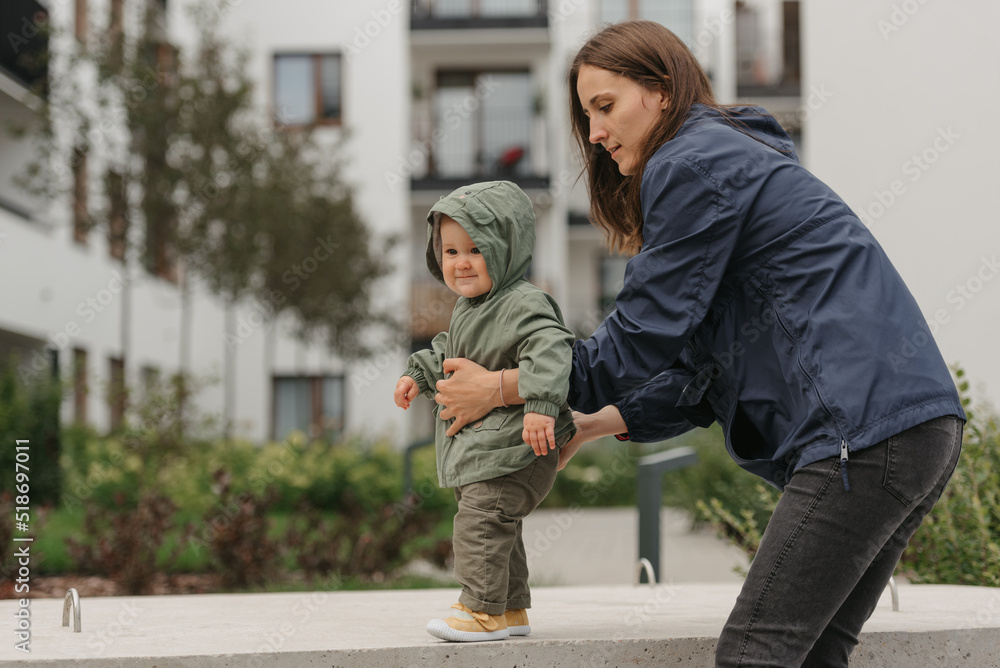 A mother is holding her pretty daughter in a raincoat on a cloudy day. A mom is teaching her female toddler how to do steps in the street at noon.