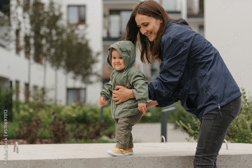 A smiling mother is holding her young daughter in a raincoat on a cloudy day. A mom is teaching her female toddler how to do steps in the street at noon.