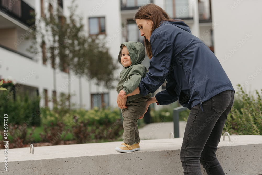 A mother is holding her young daughter in a raincoat on a cloudy day. A mom is teaching her female toddler how to do steps in the street at noon.