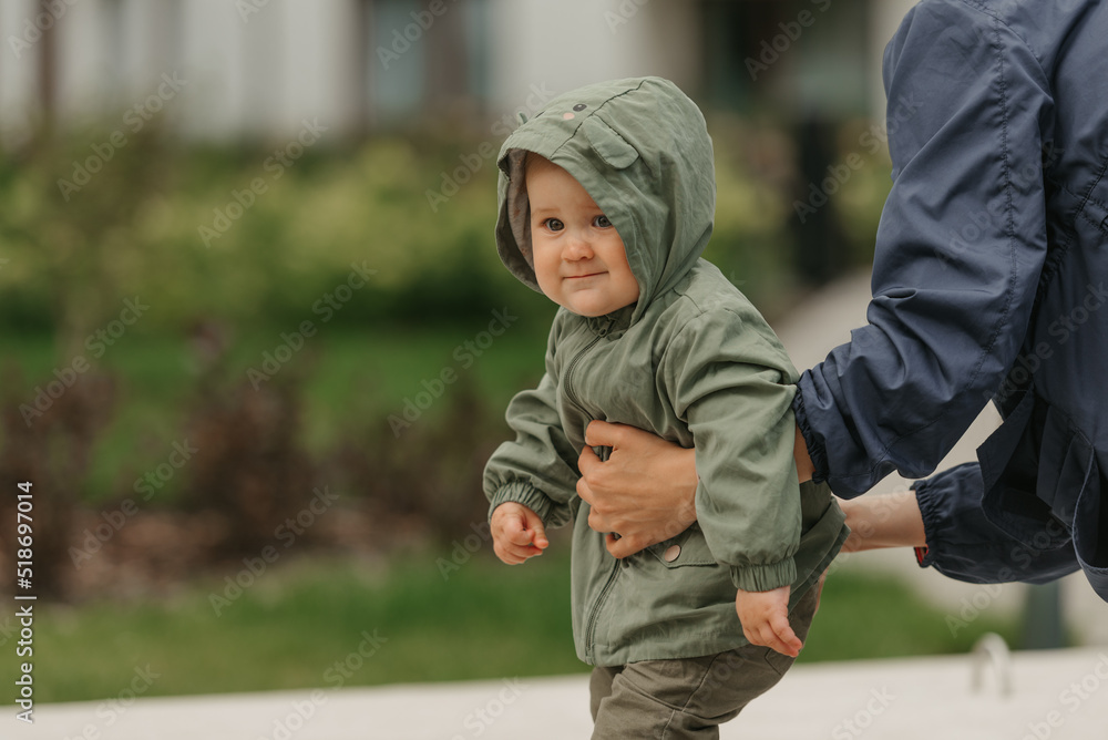 A close photo of a female toddler who is held by her mom in the street at noon. A mother is teaching her young daughter how to do steps near their home on a cloudy day.