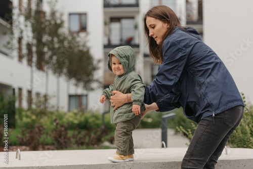 Obraz na płótnie A mother is holding her pretty daughter in a raincoat on a cloudy day. A mom is teaching her female toddler how to do steps in the street at noon. w salonie