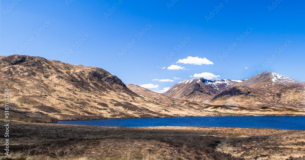 Beautiful calm loch or lake surrounded by rugged hills, brown scrub grassland and blue sky with a few clouds. Copy space. Scottish Highlands, Scotland, UK. Beautiful calm loch or lake surrounded by rugged hills, brown scrub grassland and blue sky with a few clouds. Copy space. Scottish Highlands, Scotland, UK.