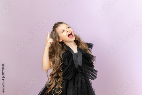 A beautiful girl points her finger up at your advertisement. Beautiful little miss with wavy hair on pink isolated background.