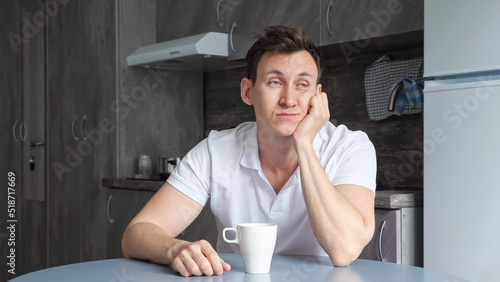 Sleepy man wants to sleep and drinks cup of coffee. Tired guy naps sitting at table in kitchen. Exhausted young father sleeps badly lately