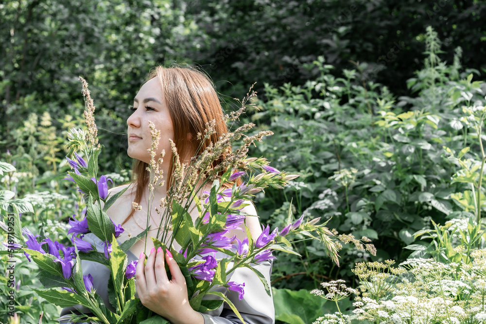 portrait of a girl in a gray satin suit among the tall grass. a model with long straight hair holds a bouquet of field herbs in the forest. the concept of femininity. portrait of a girl in a gray satin suit among the tall grass. a model with long straight hair holds a bouquet of field herbs in the forest. the concept of femininity.