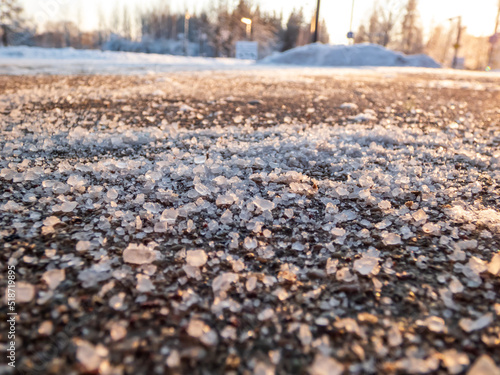 Salt grains on icy sidewalk surface in the winter. Applying salt to keep roads clear and people safe in winter weather from ice or snow. Macro view of salt grains with winter scenery in bacground photo