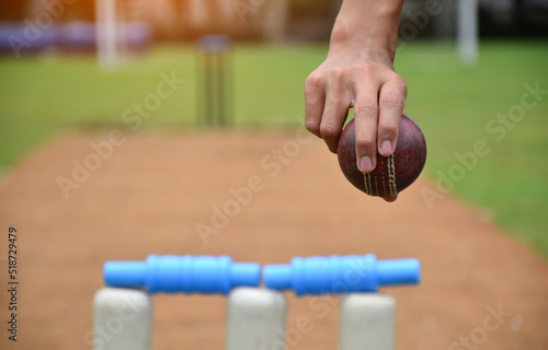 Right hand of cricketer holds old red leather cricket ball, soft and selective focus on hand, blurred cricket court background.