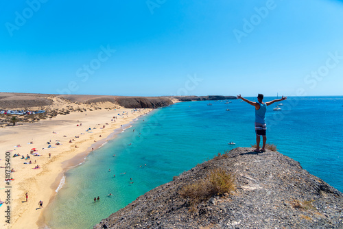 Un hombre extendiendo los brazos en una monte con el mar azul turquesa y la playa debajo. Playa Mujeres, Lanzarote. Isla canarias.